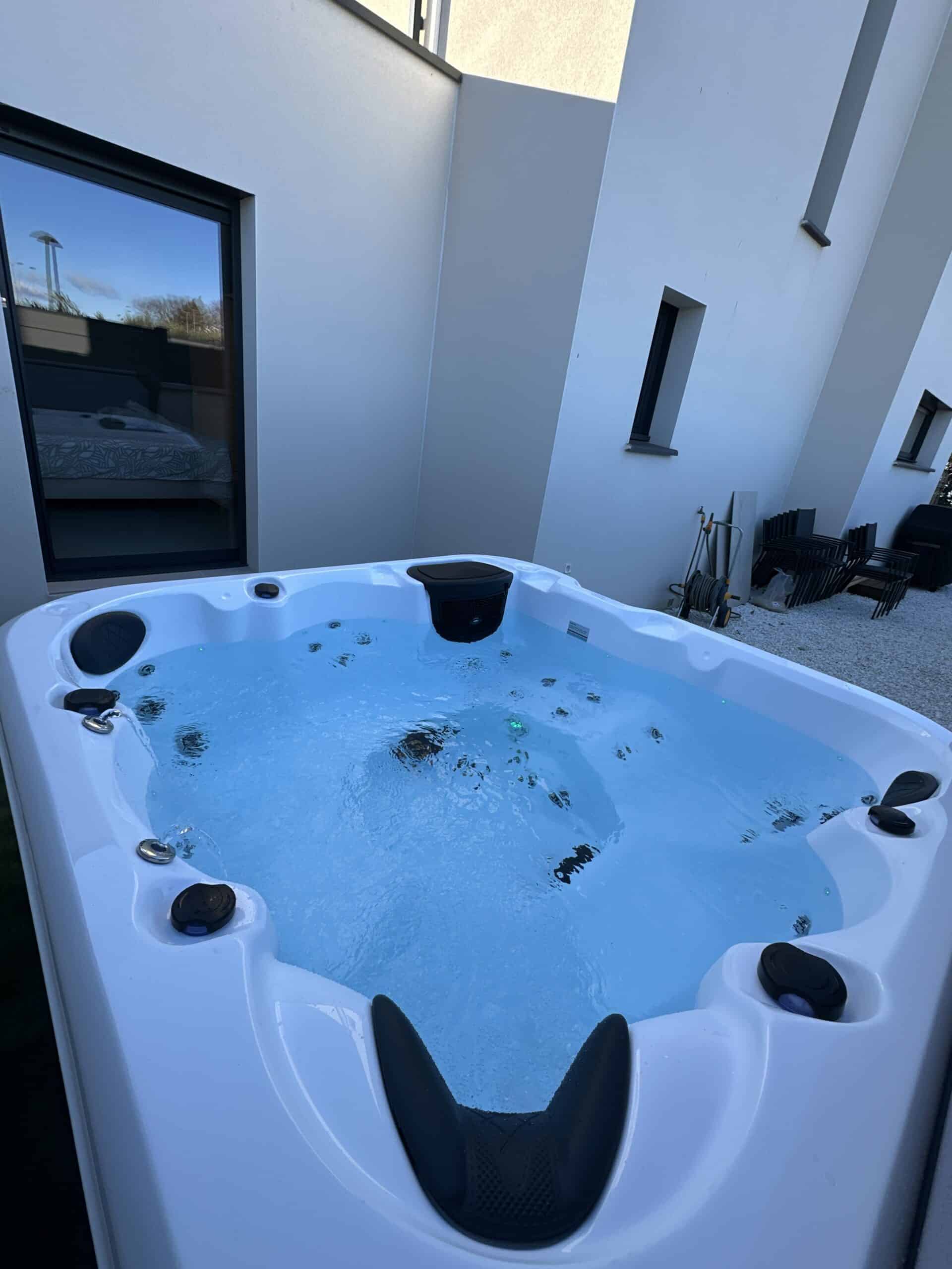Jacuzzi spa moderne blanc en extérieur, bouillonnant d'eau bleue. Reflet d'une chambre dans la fenêtre adjacente du bâtiment.