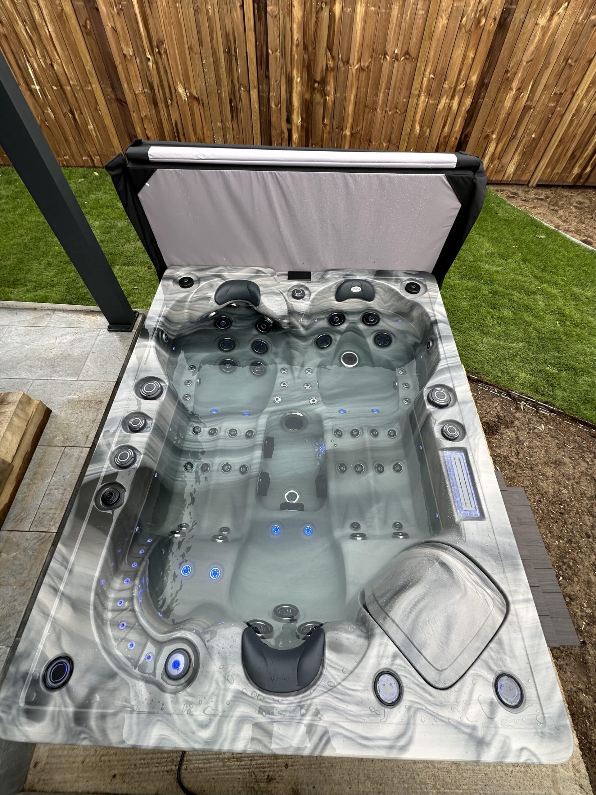 Jacuzzi spa extérieur moderne gris marbré. Bain bouillonnant avec jets et éclairage LED bleu, installé sur patio près d'une clôture en bois.