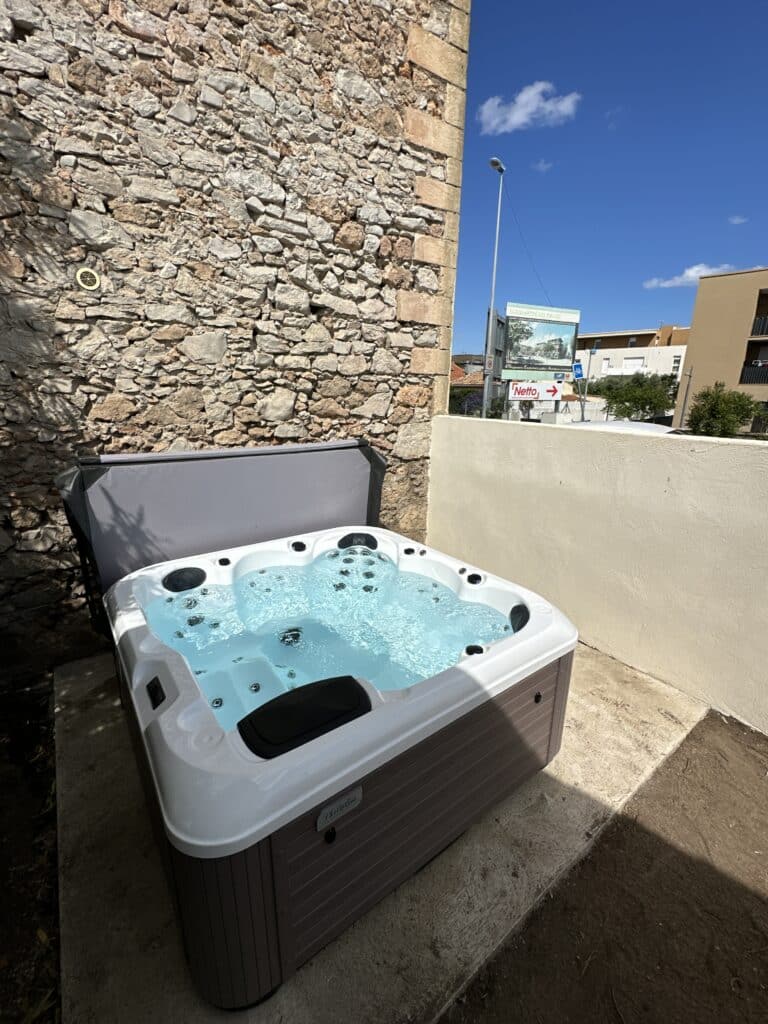 Jacuzzi extérieur bouillonnant sur une terrasse, adossé à un mur en pierre rustique sous un ciel bleu. Panneau « Netto » visible en arrière-plan.