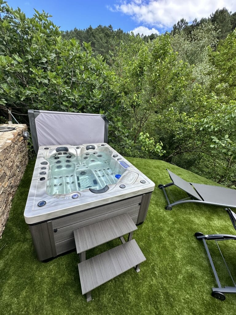 Spa extérieur moderne sur gazon, avec marches et transats, adossé à un mur de pierre. Entouré de verdure dense et d'une forêt.