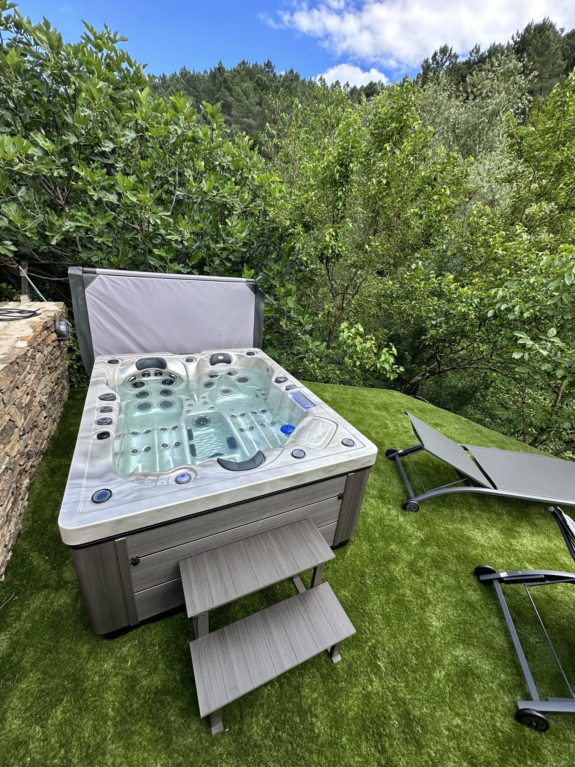 Spa extérieur moderne sur gazon, avec marches et transats, adossé à un mur de pierre. Entouré de verdure dense et d'une forêt.