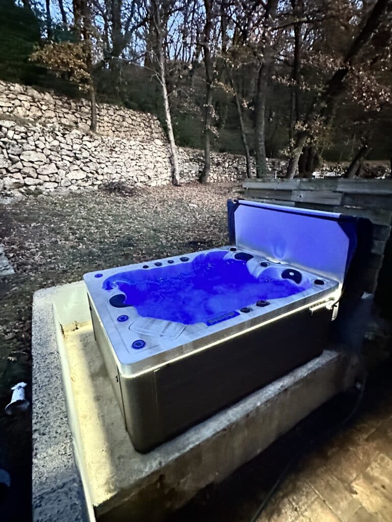 Jacuzzi Spa : Détente extérieure sous la lumière bleue. Jacuzzi extérieur au milieu de la nature la nuit. L'eau bouillonnante est éclairée en bleu vif près d'un mur de pierres sèches et d'une forêt.