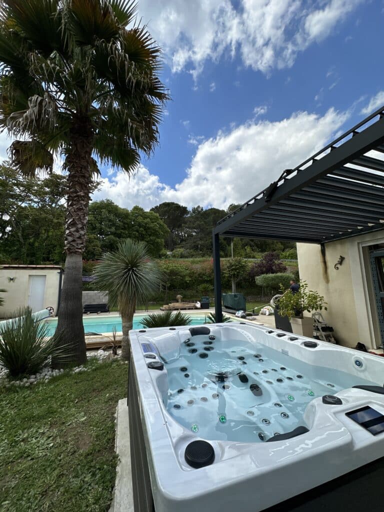 Spa extérieur blanc moderne avec eau turquoise, installé dans un jardin tropical près d'une piscine et d'une pergola bioclimatique.