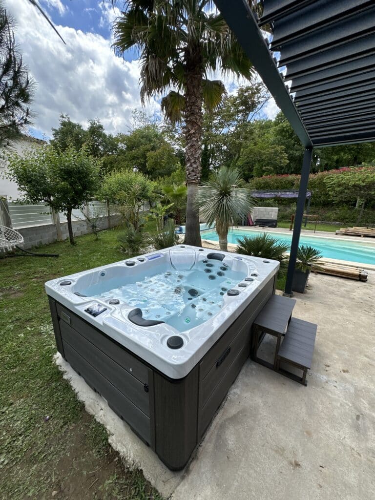 Spa jacuzzi moderne gris sur terrasse, eau bleue bouillonnante, dans un jardin tropical près d'une piscine.