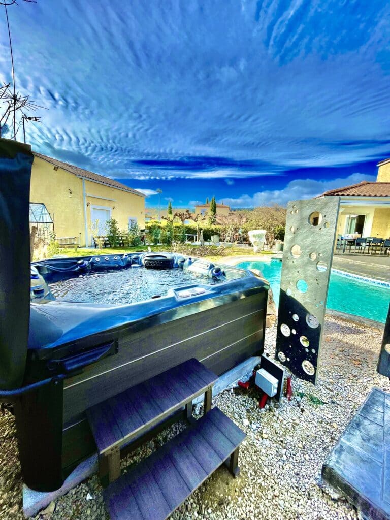 Jacuzzi extérieur moderne, piscine et ciel bleu éclatant. Spa extérieur moderne bouillonnant, marches, écran décoratif perforé, piscine et maison jaune sous un ciel bleu vif.