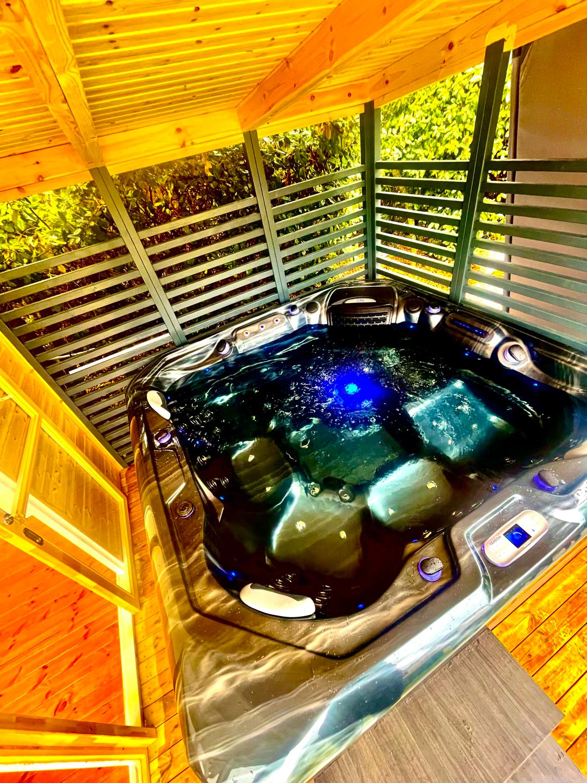Bain à remous extérieur sous abri en bois. Le jacuzzi, rempli d'eau foncée et bouillonnante, est illuminé par une vive lumière bleue.