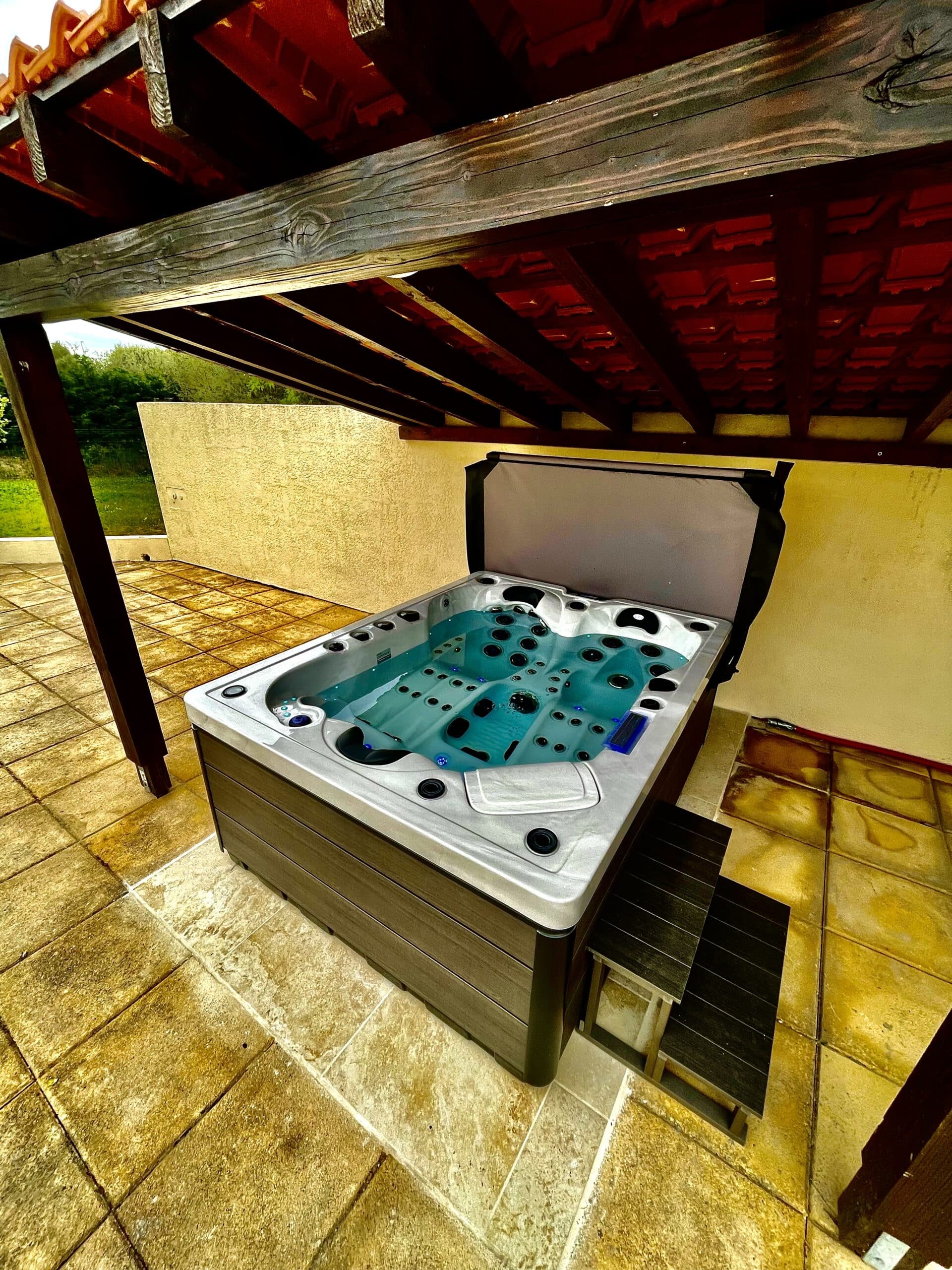 Bain à remous extérieur moderne sous une pergola en bois et tuiles. Le spa gris et l'eau turquoise reposent sur une terrasse en pierre dorée.