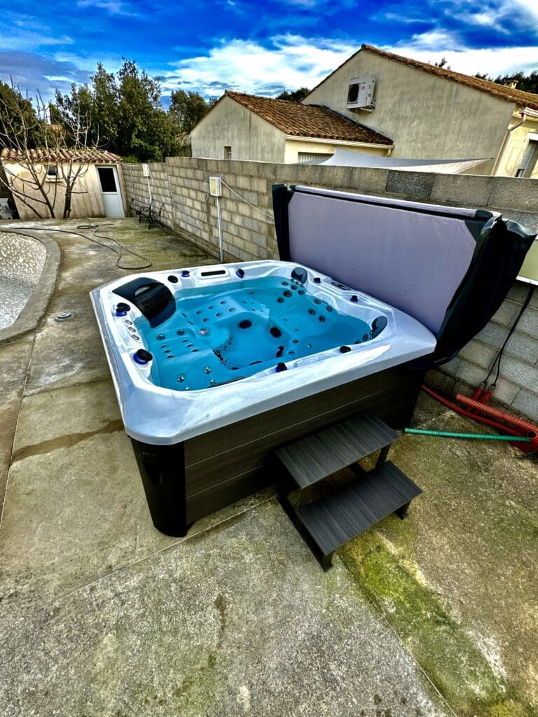 Spa Jacuzzi Extérieur : Installation sur terrasse béton Jacuzzi extérieur moderne et neuf rempli d'eau bleue. Il est posé sur une terrasse en béton, près d'un mur en blocs et d'une maison.