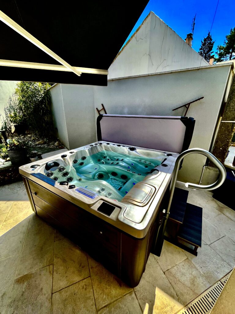 Bain à remous moderne sur terrasse, eau turquoise et carrelage beige. Couvercle ouvert, avec rampe d'accès et marches. Mur blanc et ciel bleu.