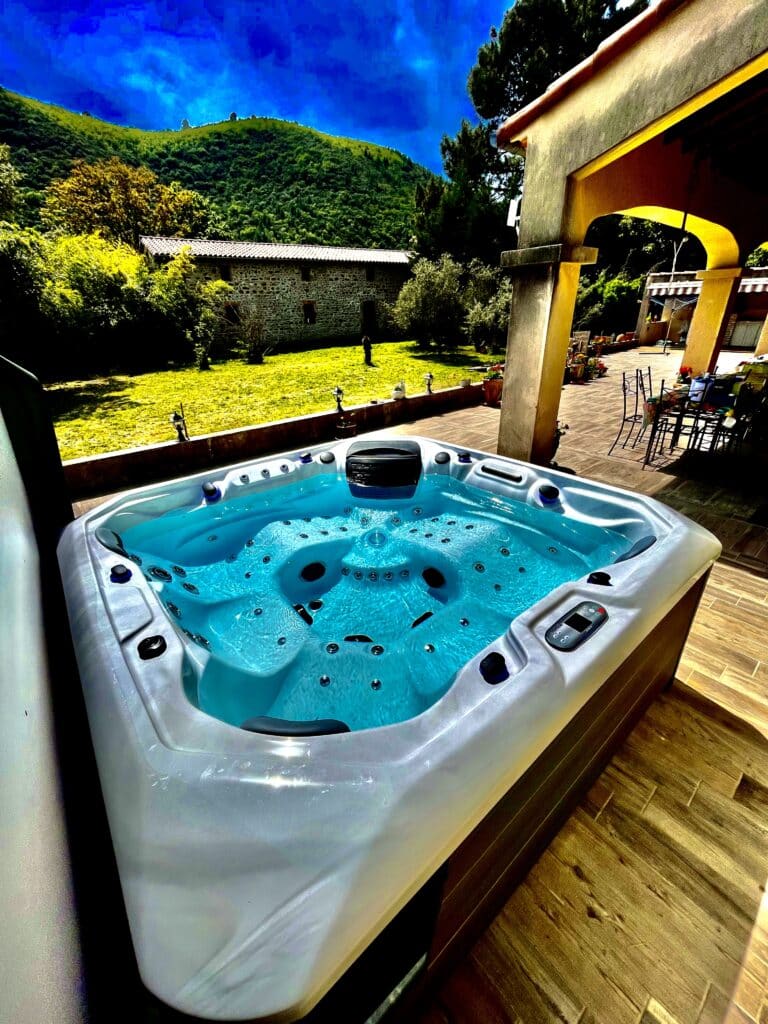 Jacuzzi extérieur moderne sur une terrasse en bois, face à un jardin ensoleillé et des collines verdoyantes sous un ciel bleu intense.