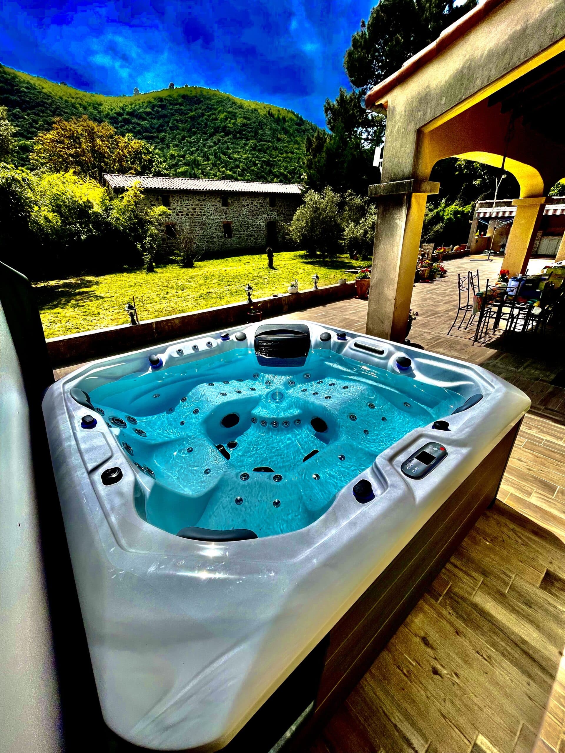 Jacuzzi extérieur moderne sur une terrasse en bois, face à un jardin ensoleillé et des collines verdoyantes sous un ciel bleu intense.