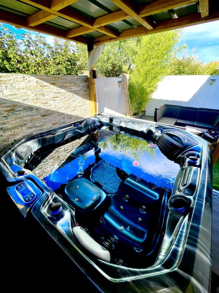 Jacuzzi bleu noir moderne sur une terrasse ensoleillée, sous pergola en bois, avec mur de pierres et verdure luxuriante. Panneau de contrôle Balboa.