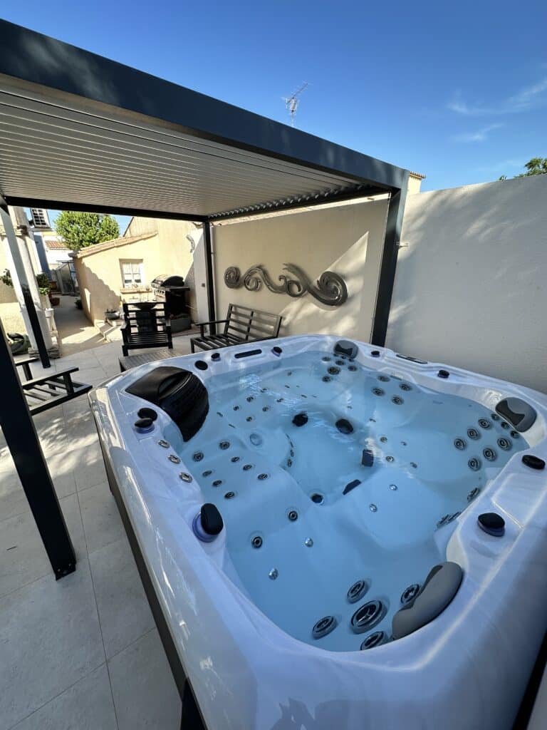 Jacuzzi spa moderne sur terrasse extérieure, protégé par une pergola. Sculpture murale ondulée et banc pour la détente estivale.