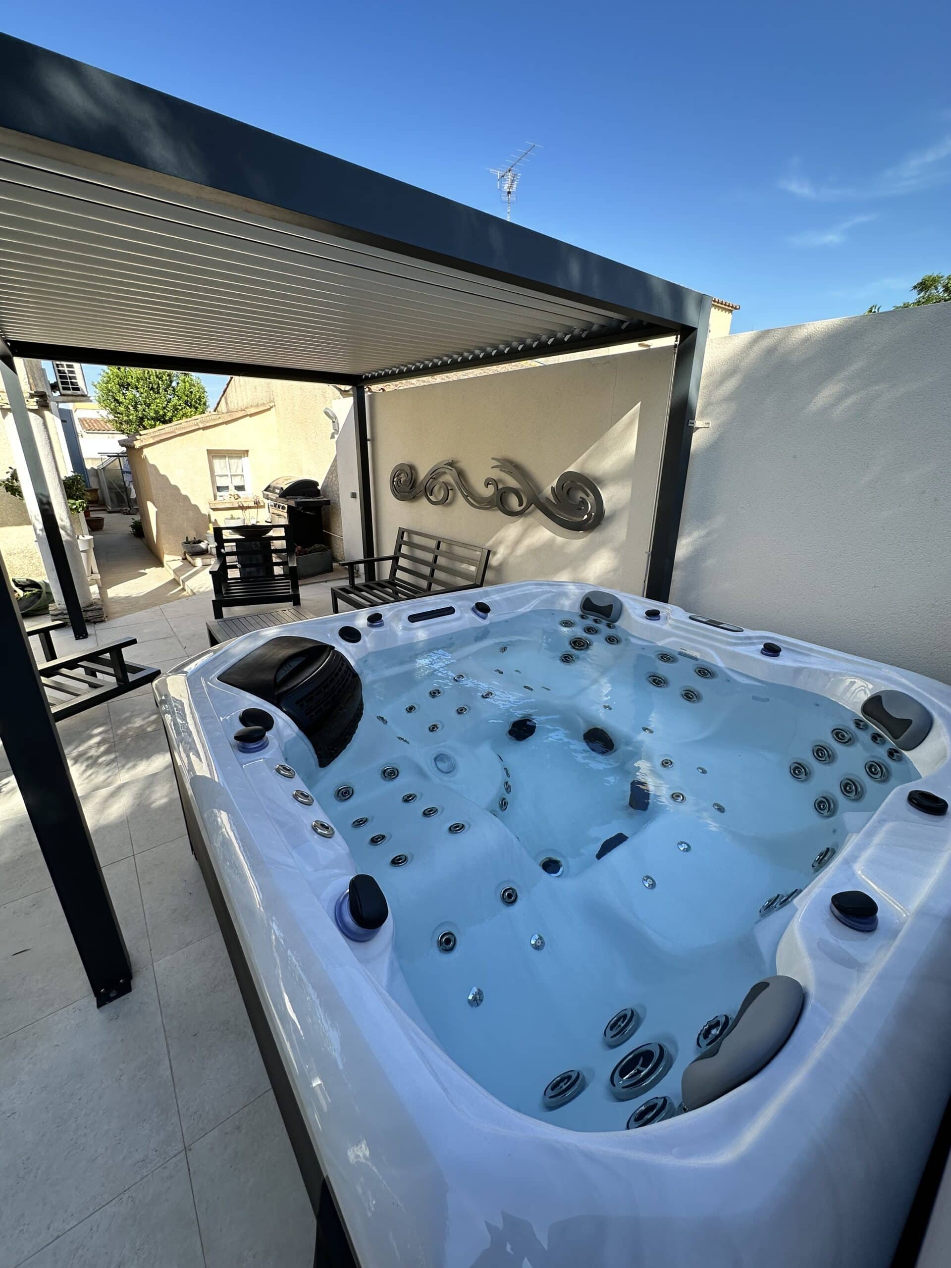 Jacuzzi spa moderne sur terrasse extérieure, protégé par une pergola. Sculpture murale ondulée et banc pour la détente estivale.