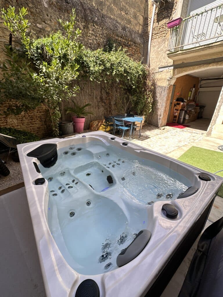Jacuzzi moderne éclairé sur une petite cour intérieure ensoleillée, murs de pierre ancienne et végétation luxuriante.
