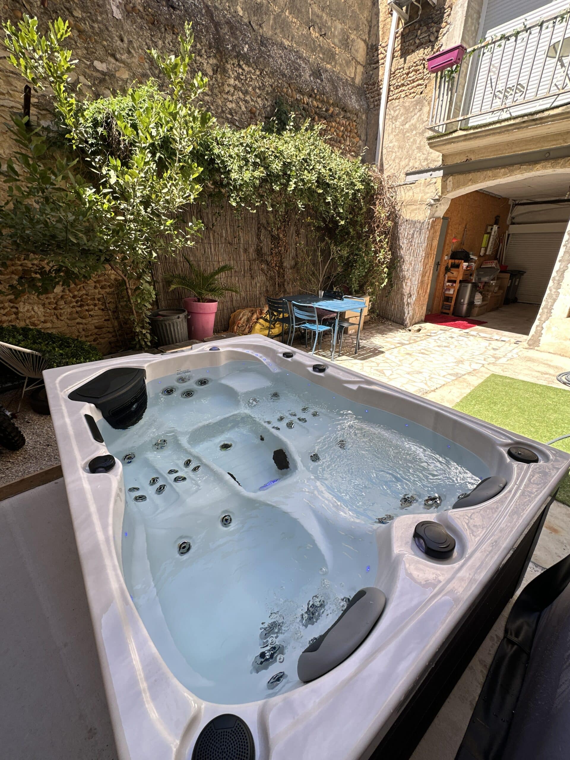 Jacuzzi moderne éclairé sur une petite cour intérieure ensoleillée, murs de pierre ancienne et végétation luxuriante.