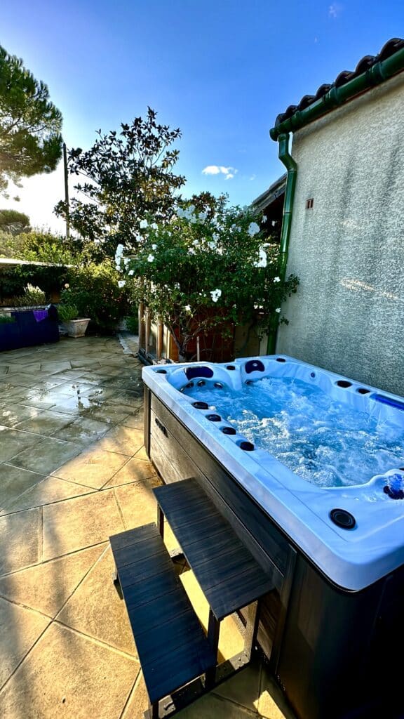 Jacuzzi spa moderne sur terrasse : l'ultime détente en extérieur. Spa extérieur bouillonnant sur une terrasse ensoleillée, entouré de buissons à fleurs blanches et d'un mur de maison. Soleil couchant.
