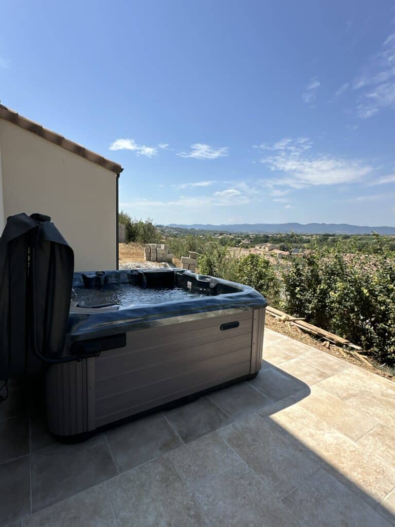 Spa jacuzzi extérieur : détente et vue panoramique. Jacuzzi spa moderne sur terrasse en carrelage beige. Vue panoramique sur la vallée, le village et les montagnes sous un ciel bleu.