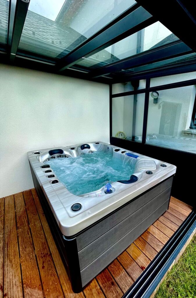 Jacuzzi extérieur privé : Détente et spa sous verrière. Jacuzzi bouillonnant sous une verrière moderne. Revêtement gris, coque argentée et terrasse en bois brun pour un moment spa extérieur.