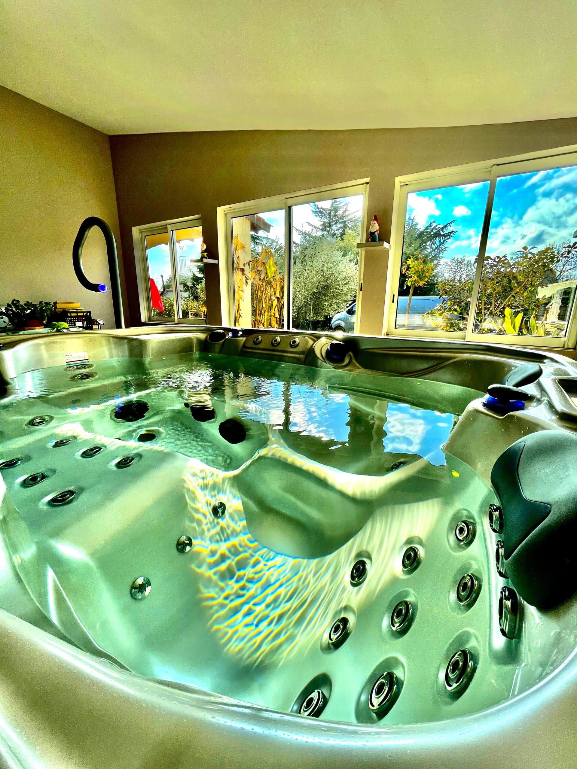 Spa intérieur lumineux; l'eau verte et agitée reflète le ciel bleu et les arbres via les baies vitrées. Jets chromés et cadre relaxant.