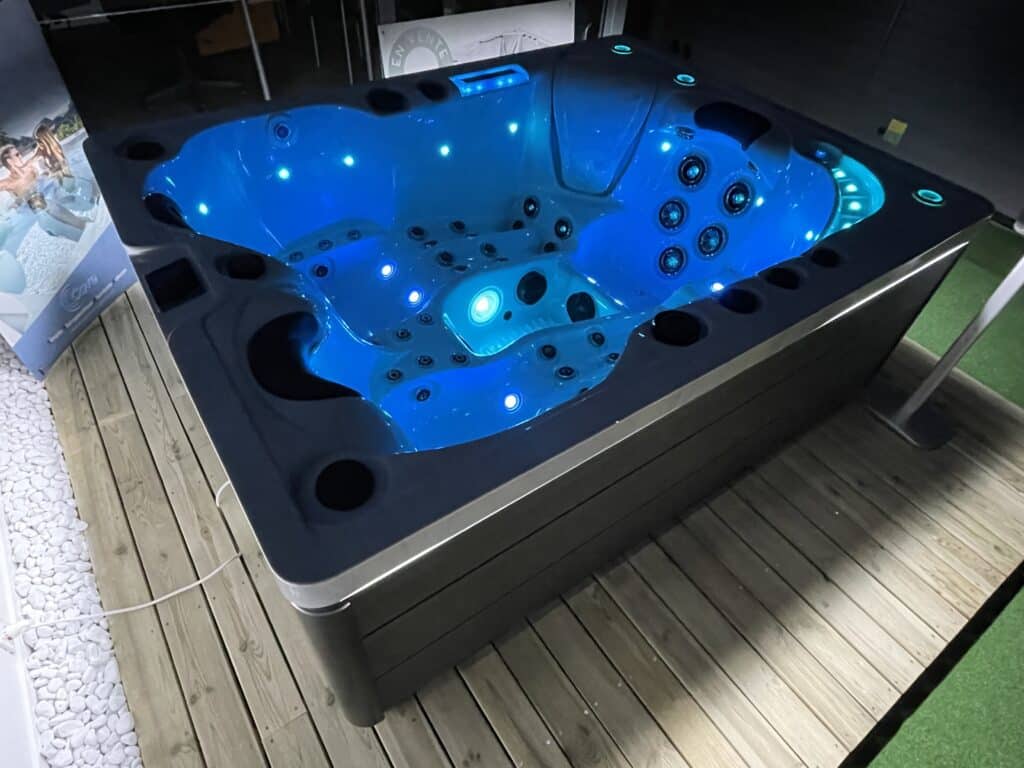 Spa Jacuzzi LED bleu éclatant | Modèle de luxe Spa Jacuzzi moderne Cove avec cuve bleue illuminée par LED. Installé sur terrasse en bois avec galets blancs. Texte "EN VENTE".