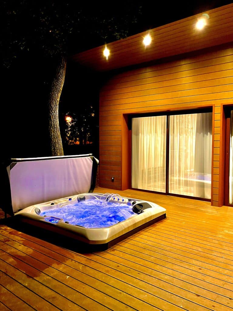Jacuzzi luxe : détente nocturne sur terrasse en bois Jacuzzi bouillonnant au LED bleu sur une terrasse en bois la nuit. Lumière chaleureuse sur la façade moderne de la maison.
