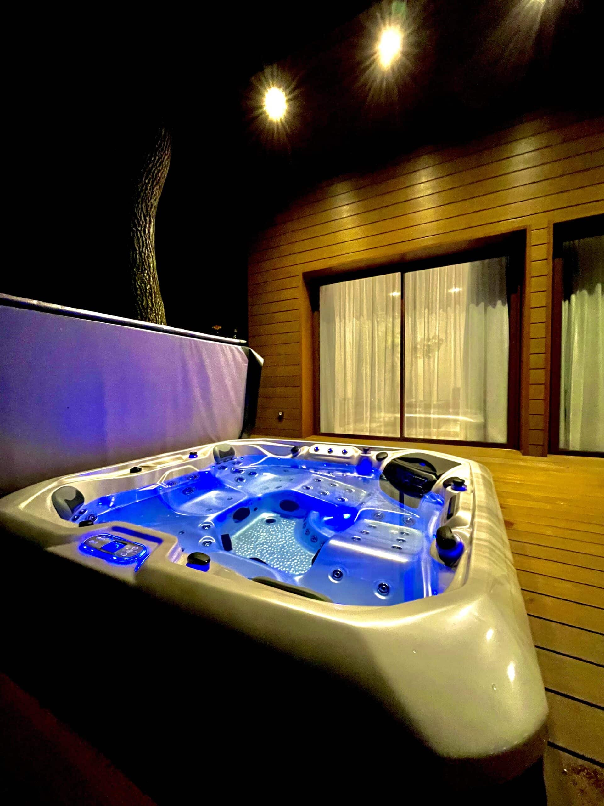Grand spa extérieur éclairé en bleu vif sur une terrasse en bois. La nuit, des spots chauds illuminent la façade moderne en lattes.