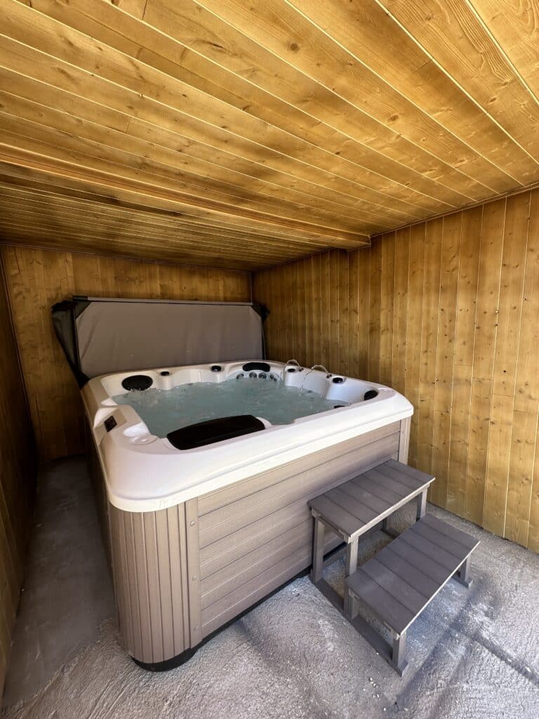 Jacuzzi et Spa Privé : Détente sous un Abri en Bois Chaleureux Jacuzzi moderne et spacieux dans un abri entièrement lambrissé de bois chaleureux. L'eau bouillonne, avec marches d'accès grises.