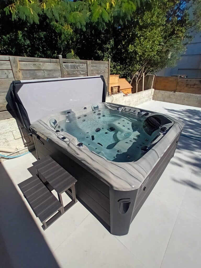 Grand jacuzzi extérieur moderne, coque marbrée, couverture ouverte, rempli d'eau bouillonnante. Escaliers sur une terrasse ensoleillée.