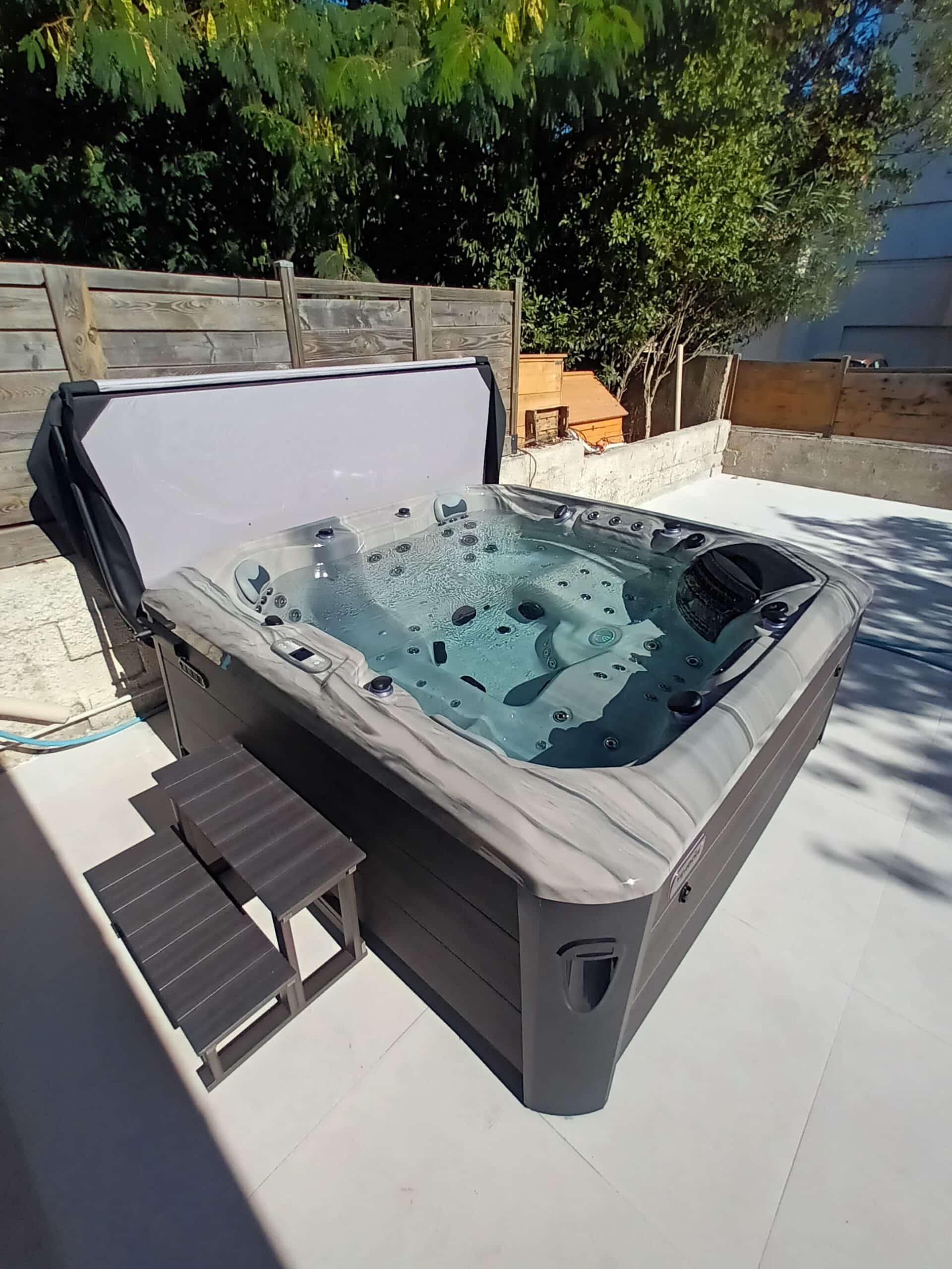 Grand jacuzzi extérieur moderne, coque marbrée, couverture ouverte, rempli d'eau bouillonnante. Escaliers sur une terrasse ensoleillée.