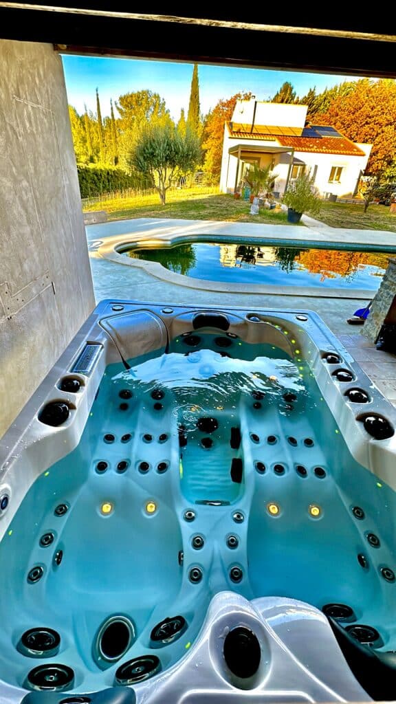 Spa jacuzzi bouillonnant bleu, vue plongeante sur une piscine extérieure. Fond : jardin ensoleillé style méditerranéen et maison.