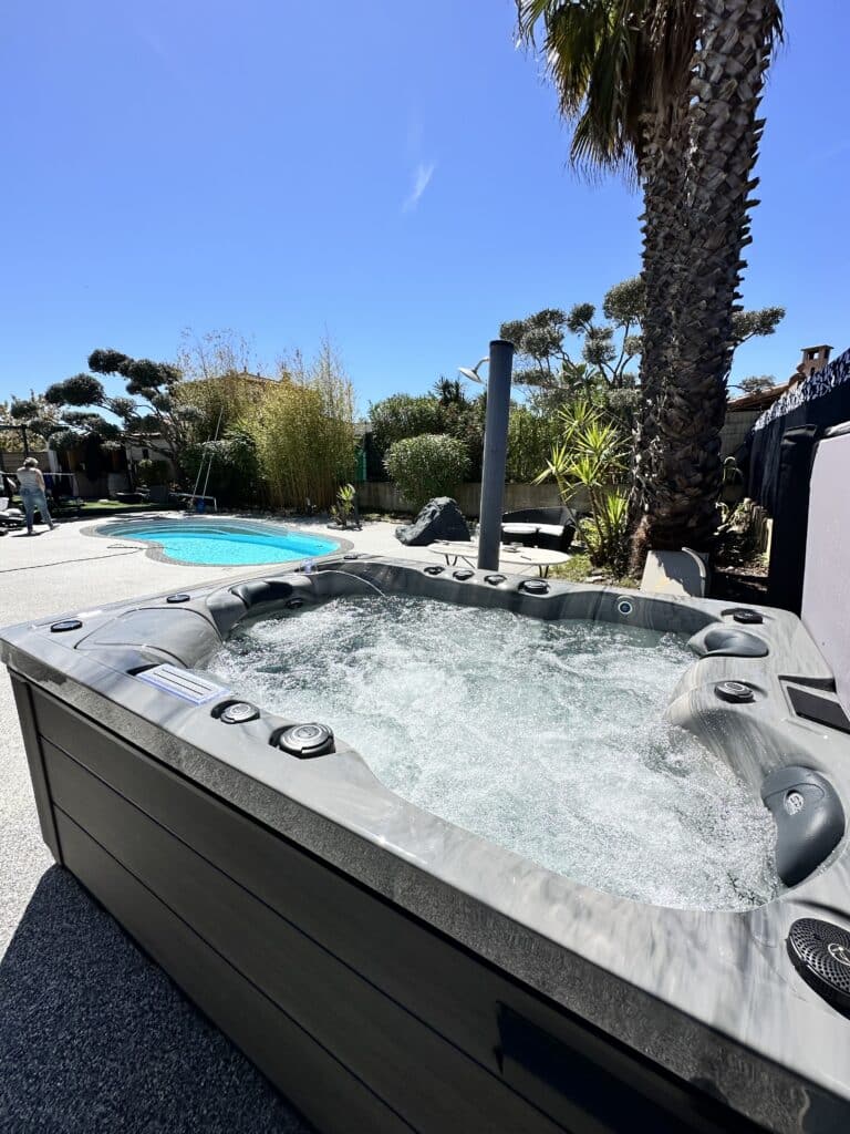 Jacuzzi Spa extérieur en action près de la piscine. Jacuzzi extérieur moderne gris avec hydrojets bouillonnants. À l'arrière-plan, piscine, palmiers et jardin luxuriant sous le soleil.