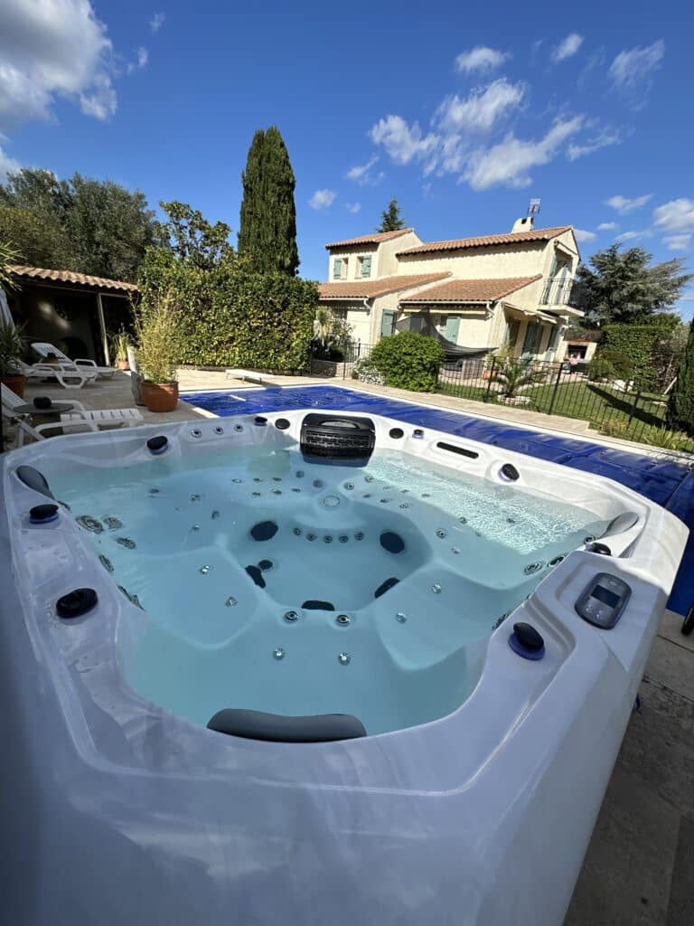 Jacuzzi blanc en extérieur, eau claire, avec un panneau de contrôle VENDÔME. Piscine couverte et maison provençale au soleil.