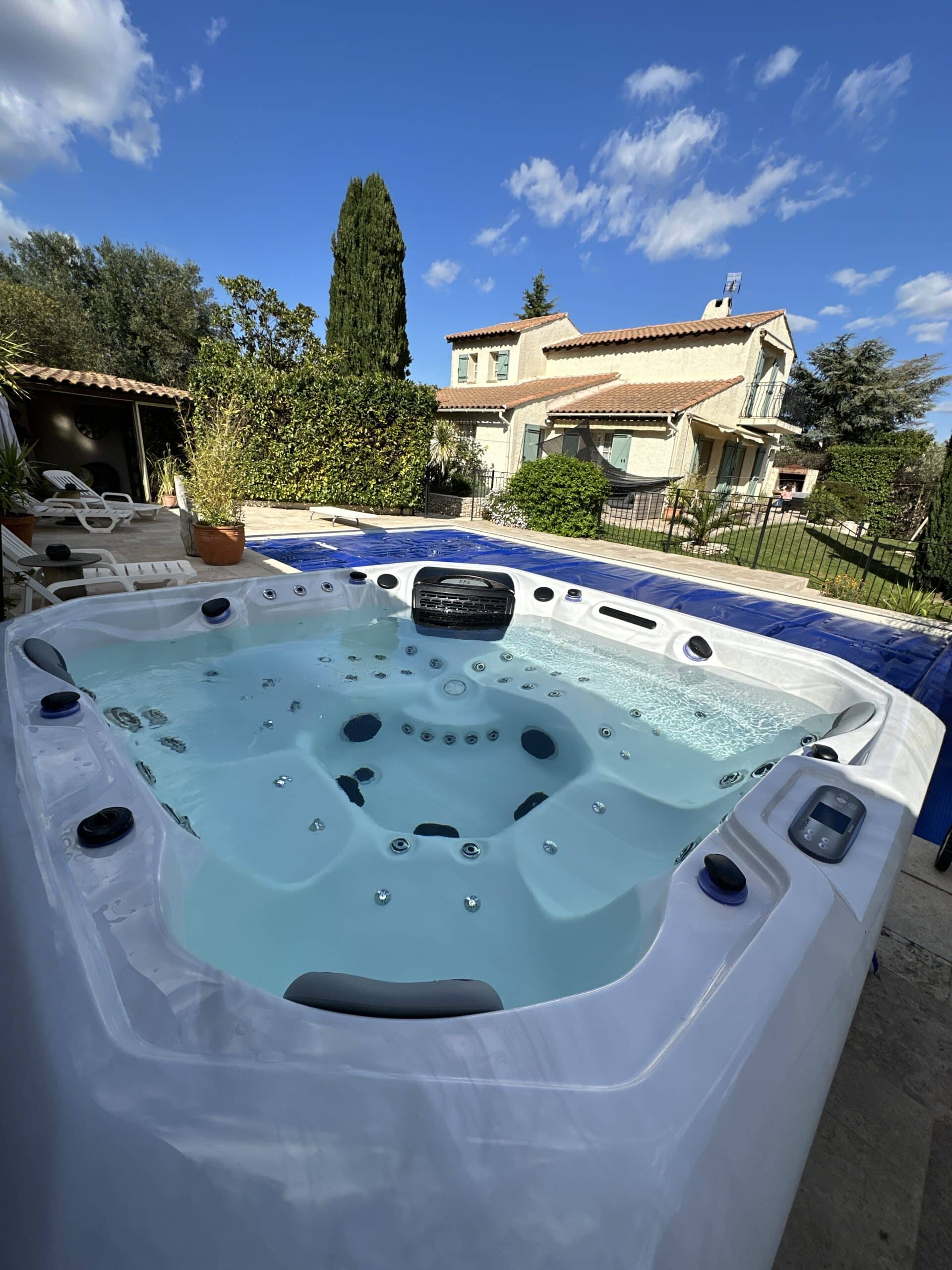 Jacuzzi blanc en extérieur, eau claire, avec un panneau de contrôle VENDÔME. Piscine couverte et maison provençale au soleil.