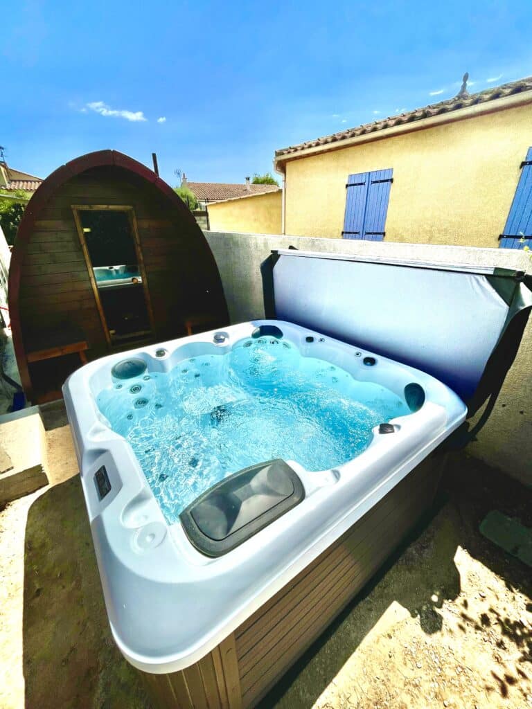 Jacuzzi bouillonnant sur une terrasse ensoleillée, avec une cabine de sauna en forme de tonneau et un mur jaune à volets bleus en arrière-plan.