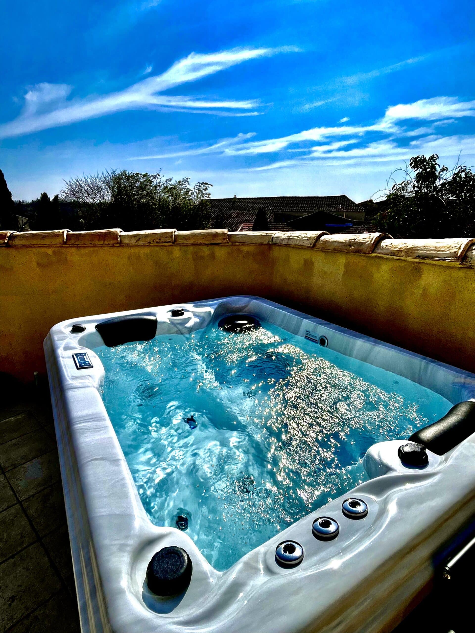 Jacuzzi blanc extérieur sur terrasse ocre. L'eau turquoise pétille sous le soleil éclatant, face à un ciel bleu azur.