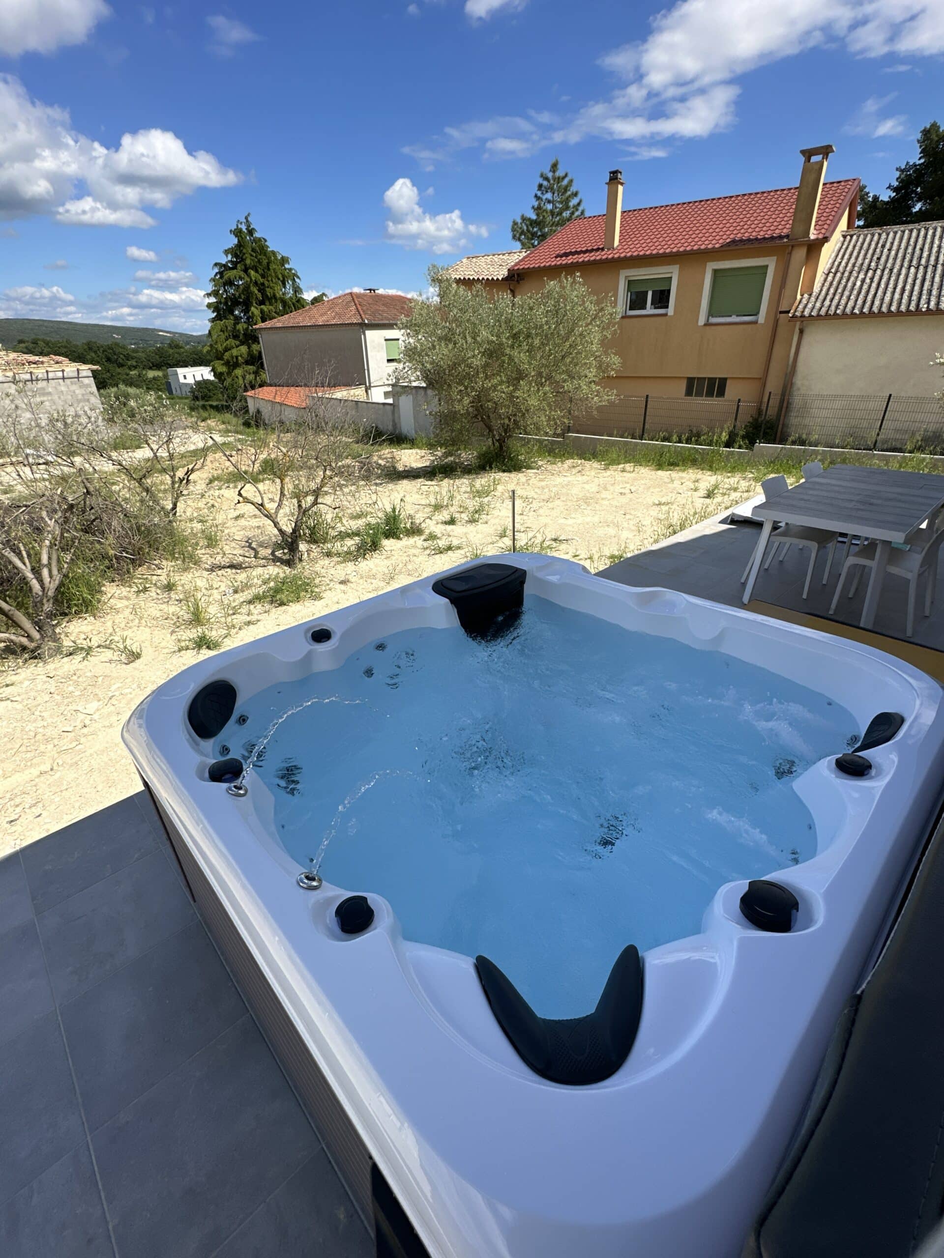 Grand spa extérieur blanc en fonctionnement sur une terrasse carrelée. Ciel bleu nuageux et jardin sec devant une maison provençale jaune.