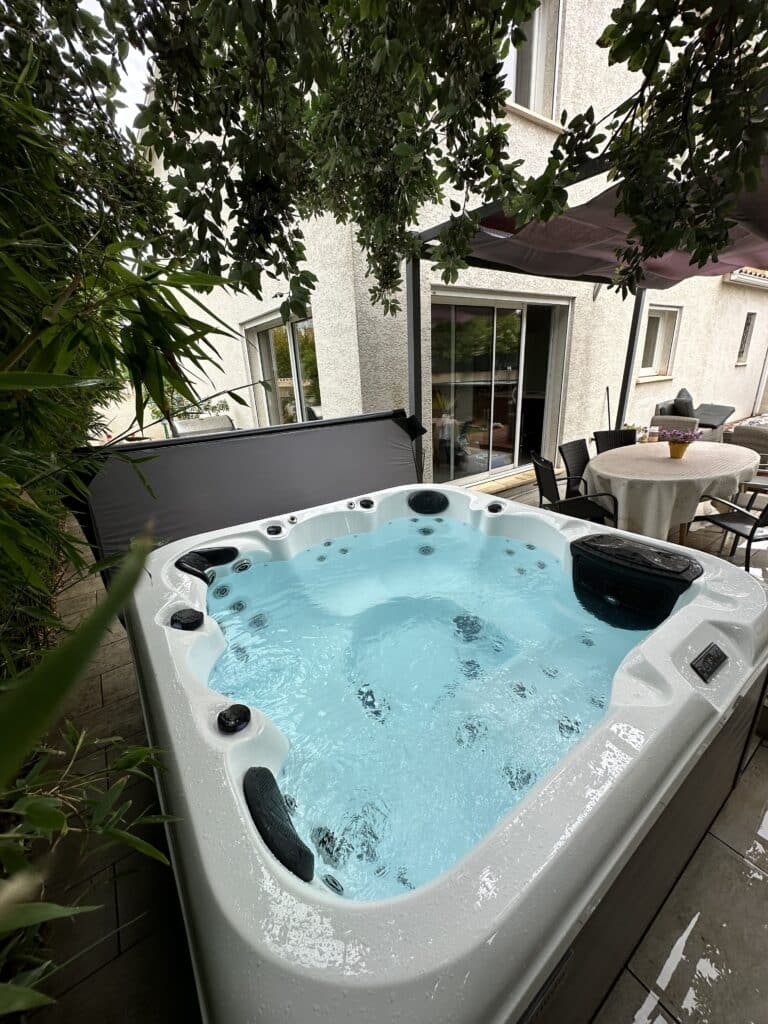 Jacuzzi spa blanc avec eau bleue bouillonnante sur une terrasse privée. Feuillage vert, façade de maison moderne et mobilier de jardin.