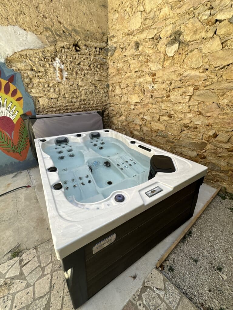 Jacuzzi extérieur Vendom : luxe et détente contre mur en pierre Spa jacuzzi extérieur Vendom en fonctionnement, eau bleue, installé dans un coin contre deux murs rustiques en vieilles pierres.