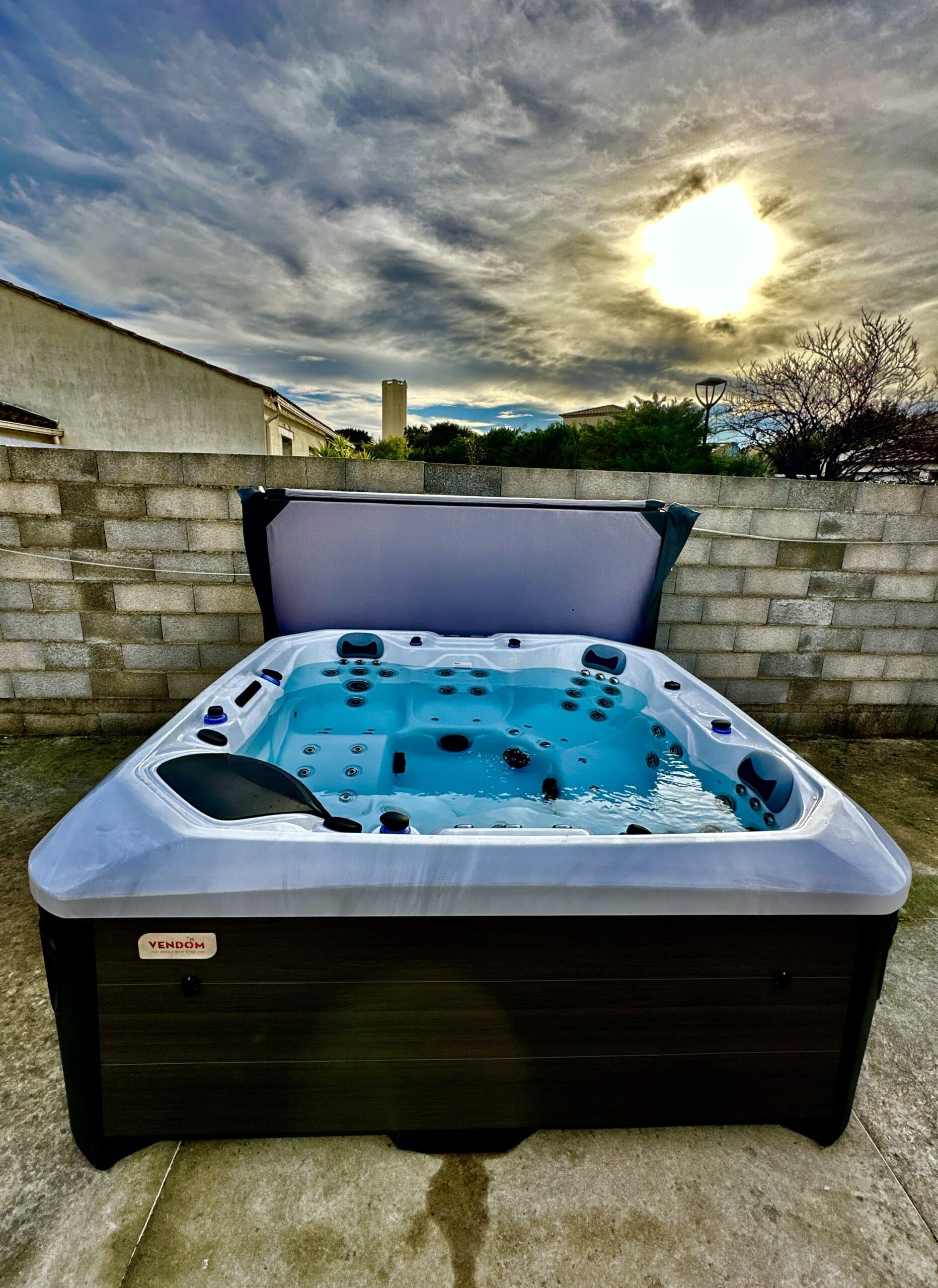 Spa jacuzzi Vendom moderne d'extérieur, eau turquoise et couvercle ouvert. Ciel dramatique du soir et soleil éclatant en arrière-plan.