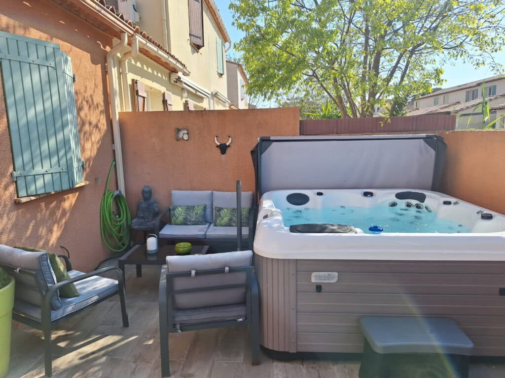 Jacuzzi VENDOM, Spa et Salon de Jardin sur Terrasse Jacuzzi VENDOM bouillonnant sur une terrasse en bois, jouxtant un salon de jardin gris. Mur extérieur orange décoré d'un Bouddha et d'une tête de taureau.