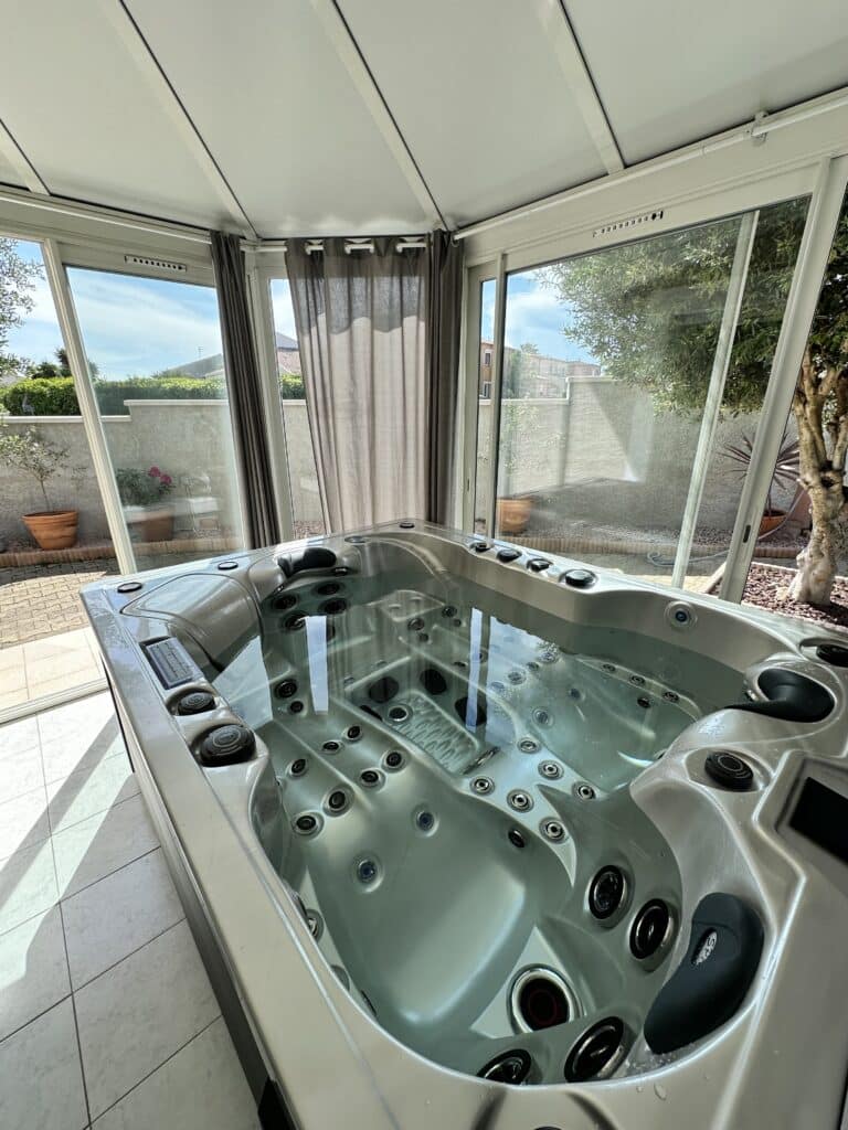 Spa Jacuzzi intérieur-extérieur lumineux Spa jacuzzi moderne gris dans une véranda lumineuse. Grandes baies vitrées ouvertes sur un jardin privé et un mur végétal sous le soleil.