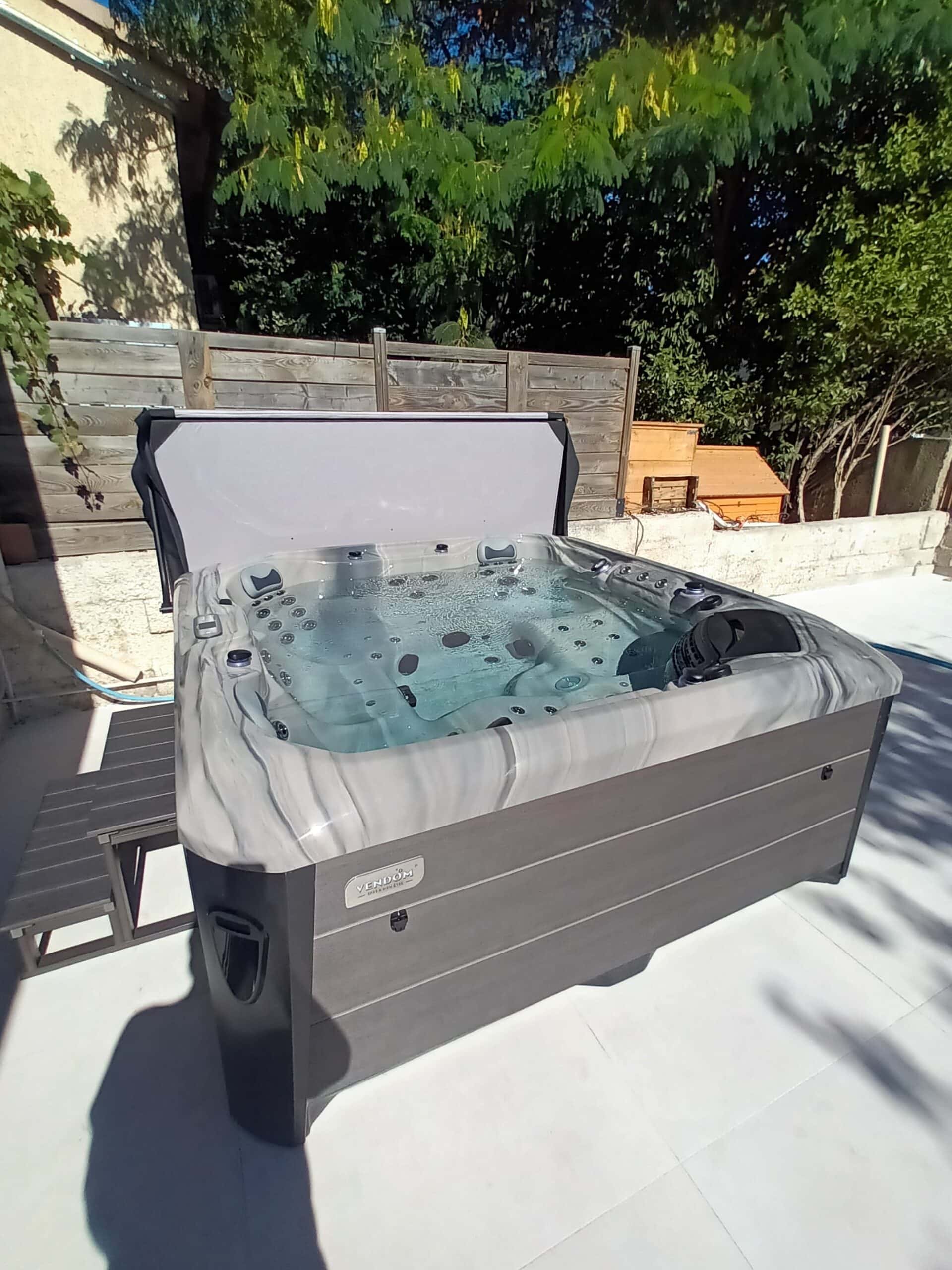 Spa Vendom gris marbré sur terrasse extérieure, jupe composite foncée, eau bouillonnante, couvercle relevé. Clôture bois en arrière-plan.