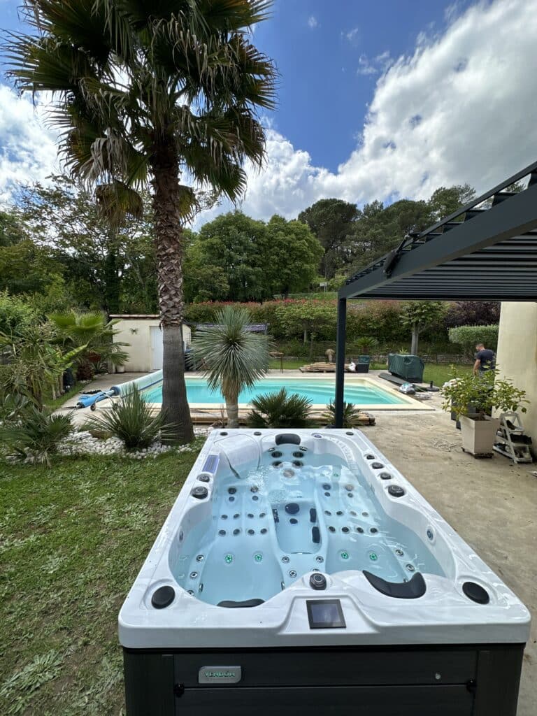 Spa jacuzzi VENDOM blanc moderne sur terrasse. Piscine et jardin luxuriant avec palmiers sous un ciel bleu ensoleillé.