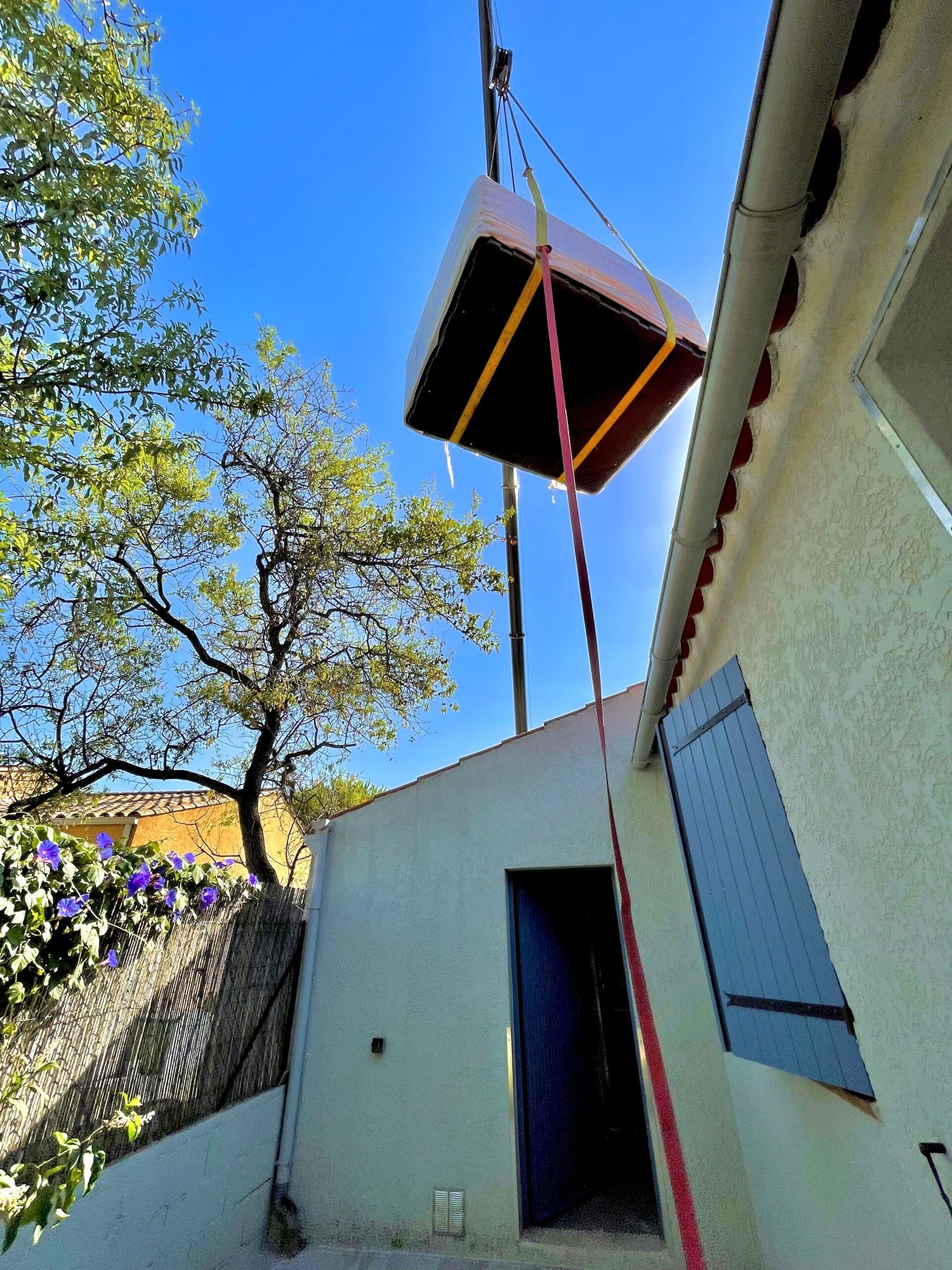 Levage d'un spa par une grue au-dessus d'une dépendance sous un ciel bleu. Installation en zone difficile d'accès près d'une façade.