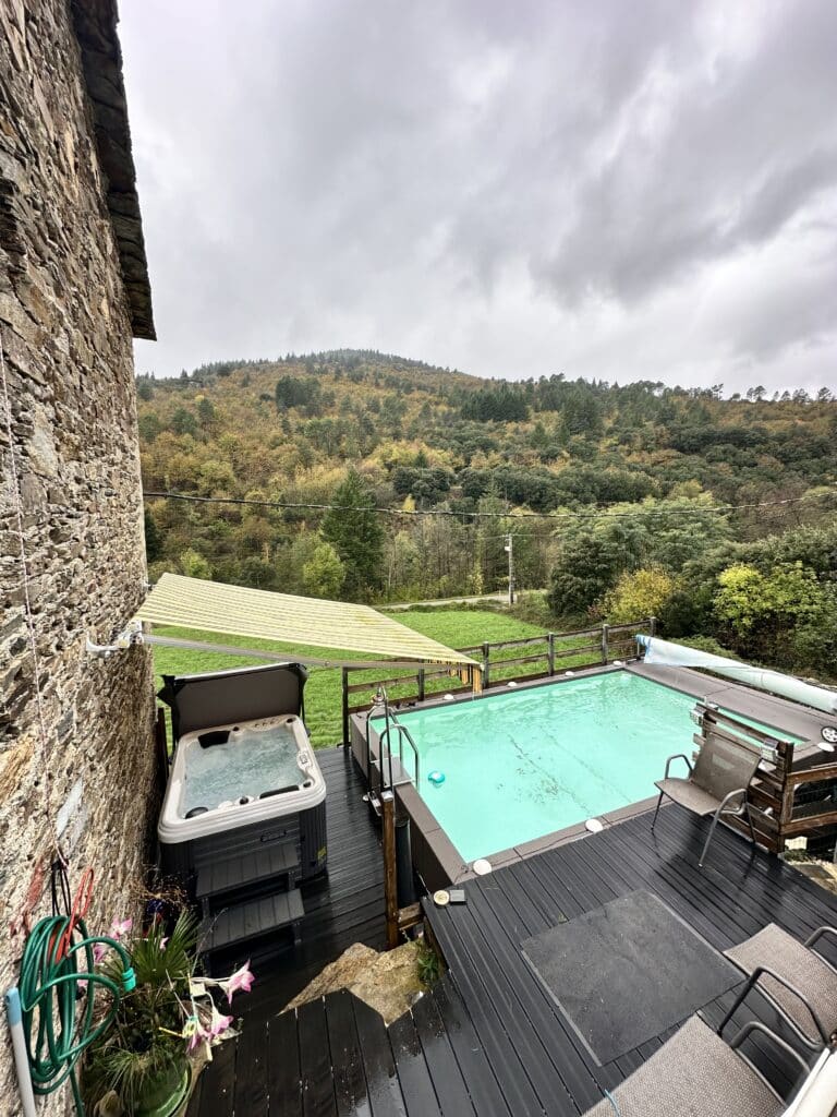 Piscine et Jacuzzi avec vue montagne sur terrasse bois Piscine et jacuzzi sur terrasse en bois noir, près d'un mur de pierre rustique. Paysage de collines d'automne sous un ciel gris.