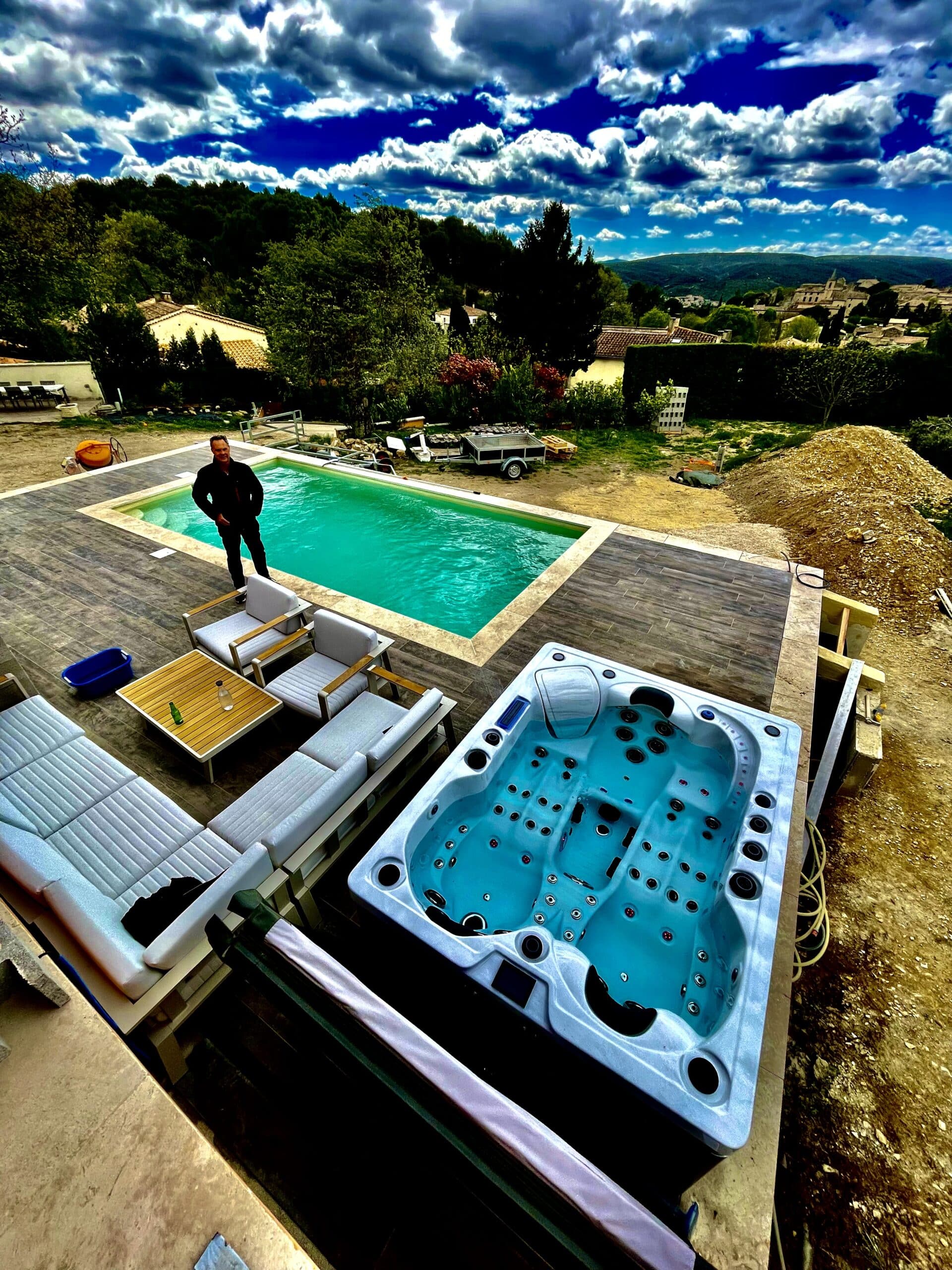 Piscine turquoise et grand spa neufs sur terrasse en bois carrelé. Homme en veste noire supervisant la construction sous un ciel dramatique.