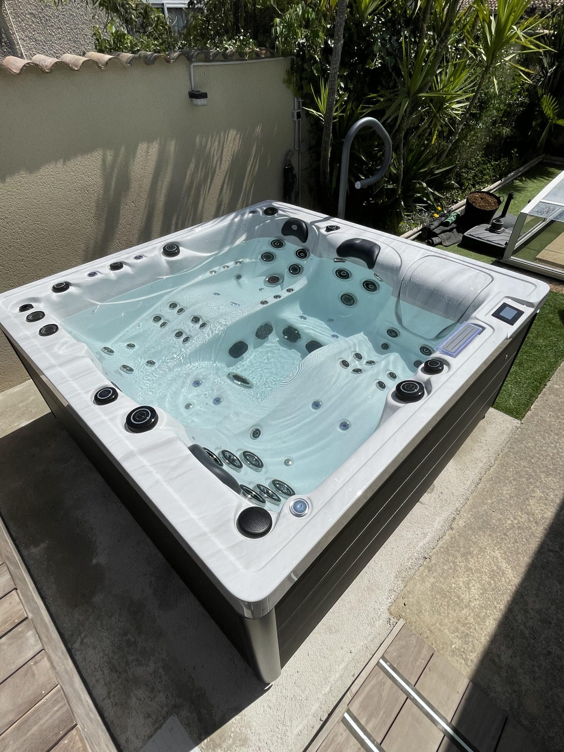 Spa balnéo carré moderne (blanc/gris) rempli d'eau claire sur une terrasse ensoleillée, près d'un mur et de végétation luxuriante.