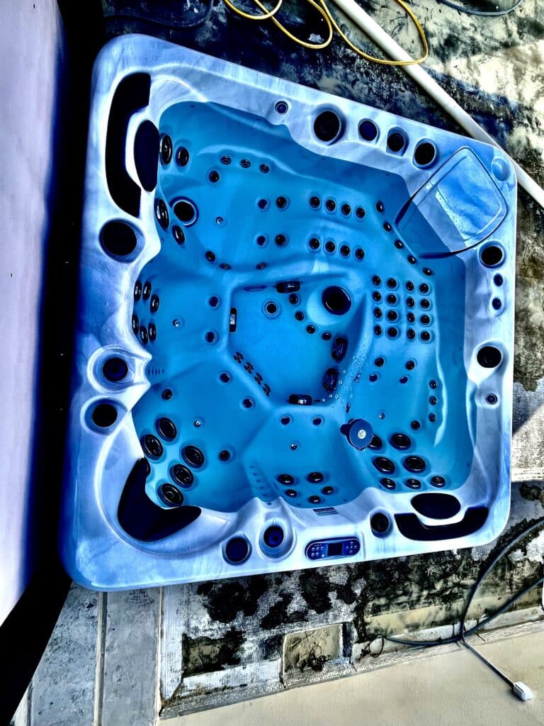 Spa rectangulaire bleu vide avec des dizaines de jets pour hydrothérapie et panneau de commande numérique, en attente d'installation sur sol brut.