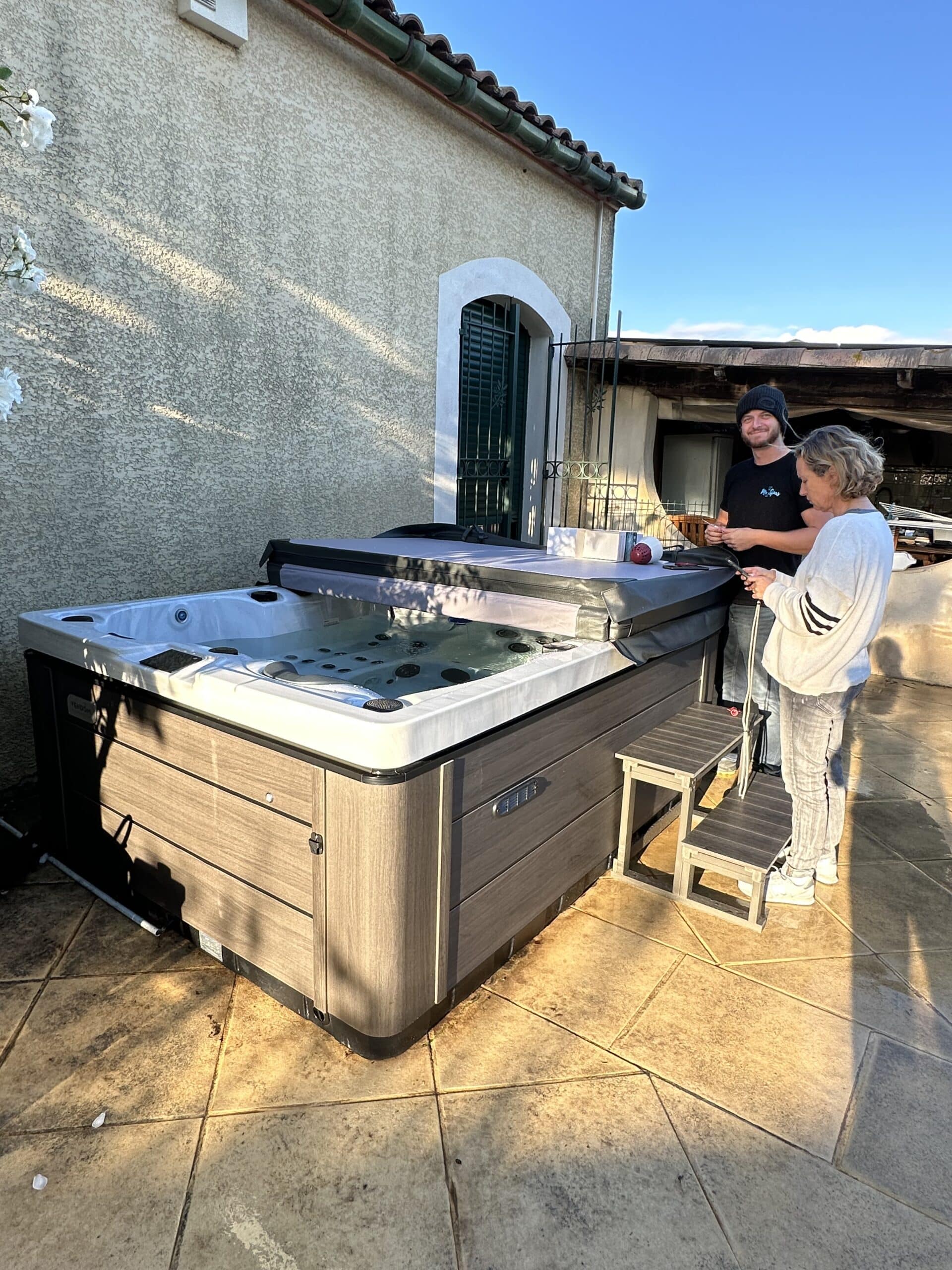 Un homme et une femme installent un grand spa extérieur moderne sur une terrasse carrelée. Le technicien donne des conseils de mise en service.